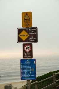 Multiple beach safety signs on a pole near the shore, warning visitors about rip currents and surf hazards.